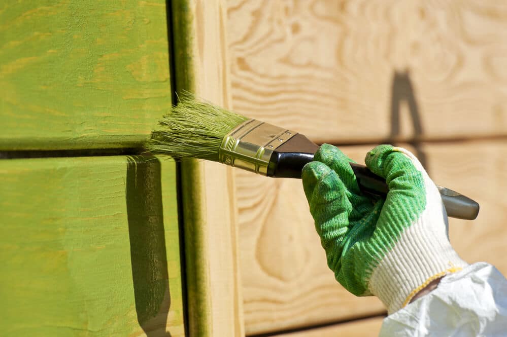 A gloved hand brushing green paint along wood cladding