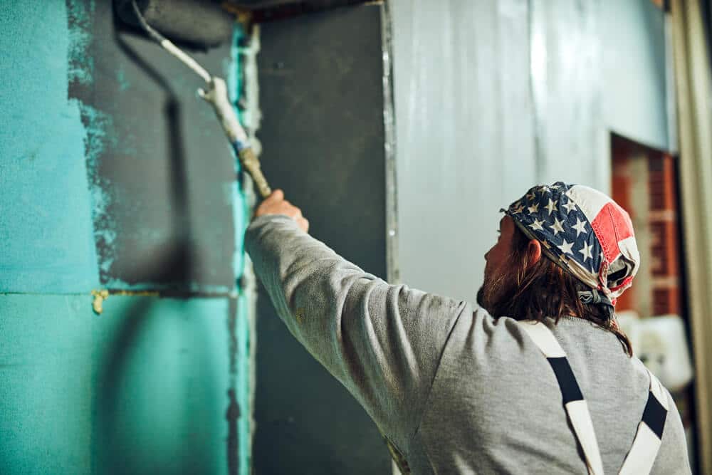 A painter rolling a teal finish onto an interior wall, seen from behind