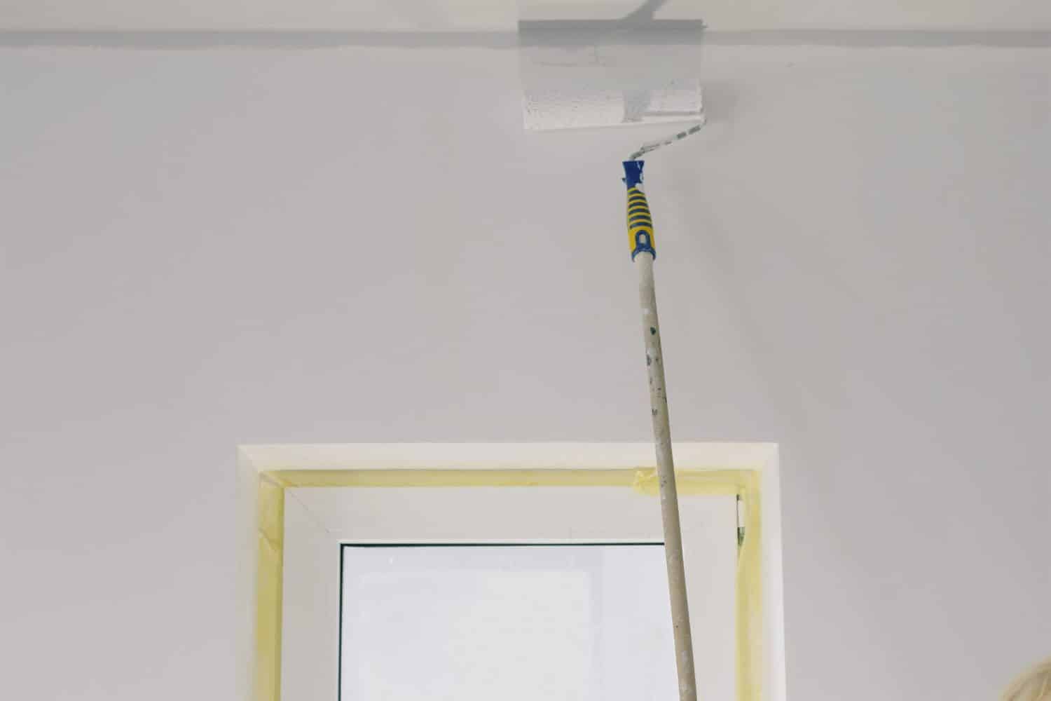 A roller on an extension pole coating a wall beside a window frame