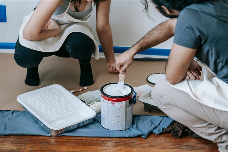 Two people kneeling beside a paint tray and an open can of paint