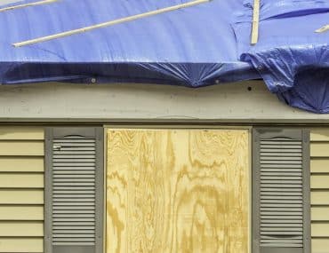 Blue tarp over a storm-damaged roof above a boarded window on sided house